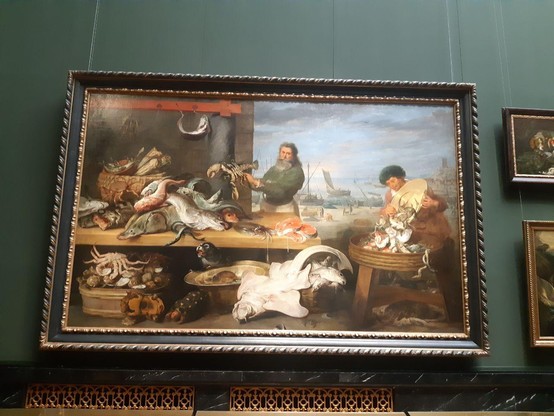 Großes Gemälde an einer grünen Wand. In der unteren Bildhälfte in Körben und auf einem Tisch verschiedene Meeresbewohner. In der Mitte und rechts jeweils Männer, die diese Fische, Krebse, Muscheln, etc. auf dem Fischmarkt anbieten. Im Hintergrund Teil eines Hafens und bewölkter Himmel
