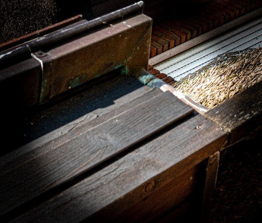 This photograph shows a close-up of part of a house facade. In the foreground, wooden boards with visible grain and small nails are visible. Above them is a metal gutter that looks aged and has a greenish oxidation patina. On the right side of the image, a section of roof made of tightly packed thatch is visible. In the background, a section of brick wall and an exterior blind are also visible. The lighting from a lamp creates strong contrasts, with some areas brightly lit and others in shadow. The overall atmosphere is rustic and weathered.