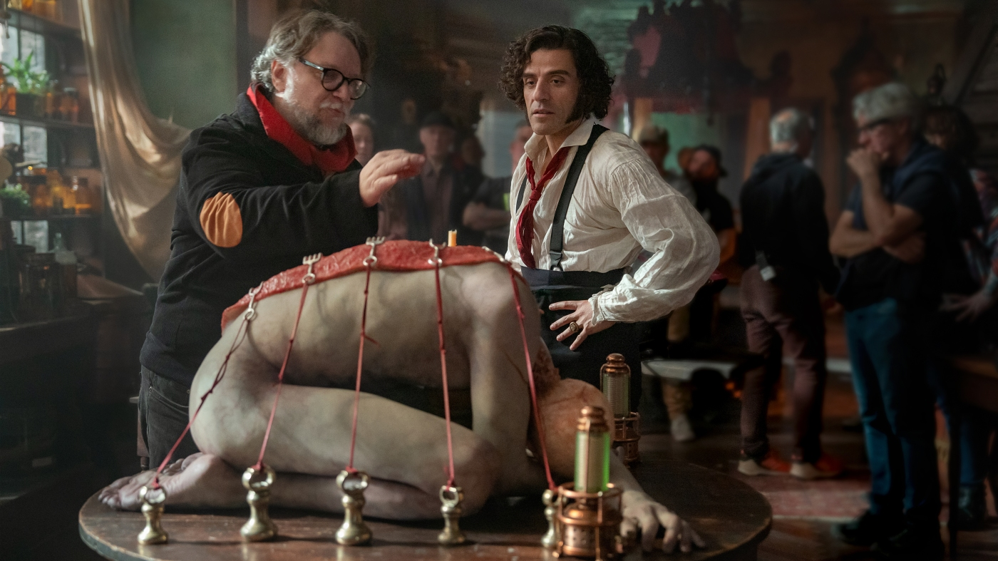Image of Guillermo del Toro and Oscar Isaac on the set of "Frankenstein." Del Toro is putting finishing touches to what appears to be a model of Frankenstein's monster, while Isaac looks on.