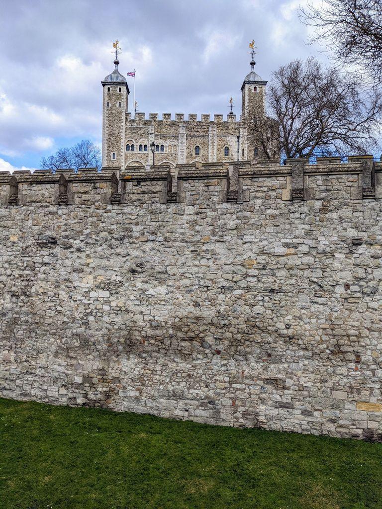 A view of the White Tower at the Tower of London, seen from behind a massive, ancient gray stone defensive wall. The stone towers are topped with golden weather vanes, and a Union Jack flag flies from a central pole against a cloudy sky.