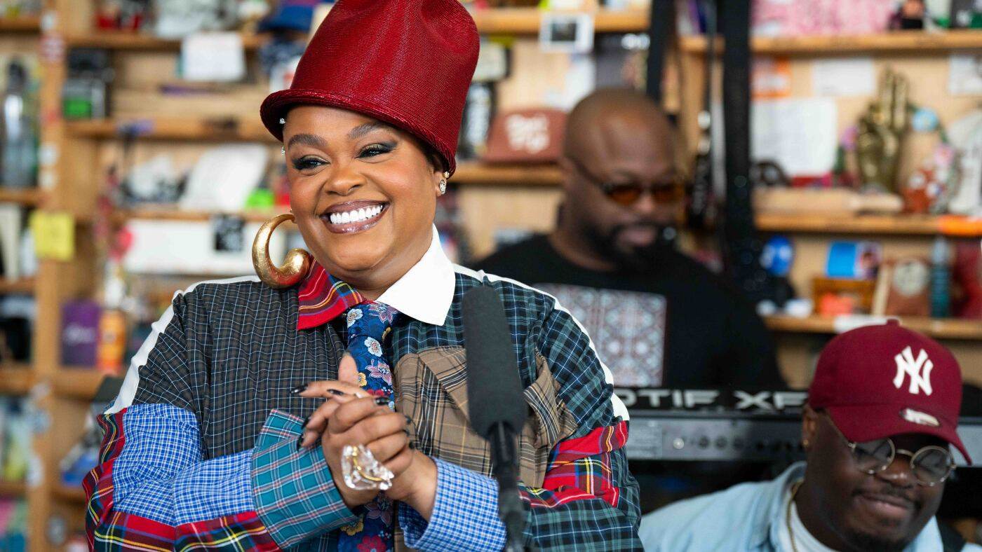 Image of Jill Scott, a Black woman wearing a red hat. She is with two Black men and and all three are in the Tiny Desk music studio.