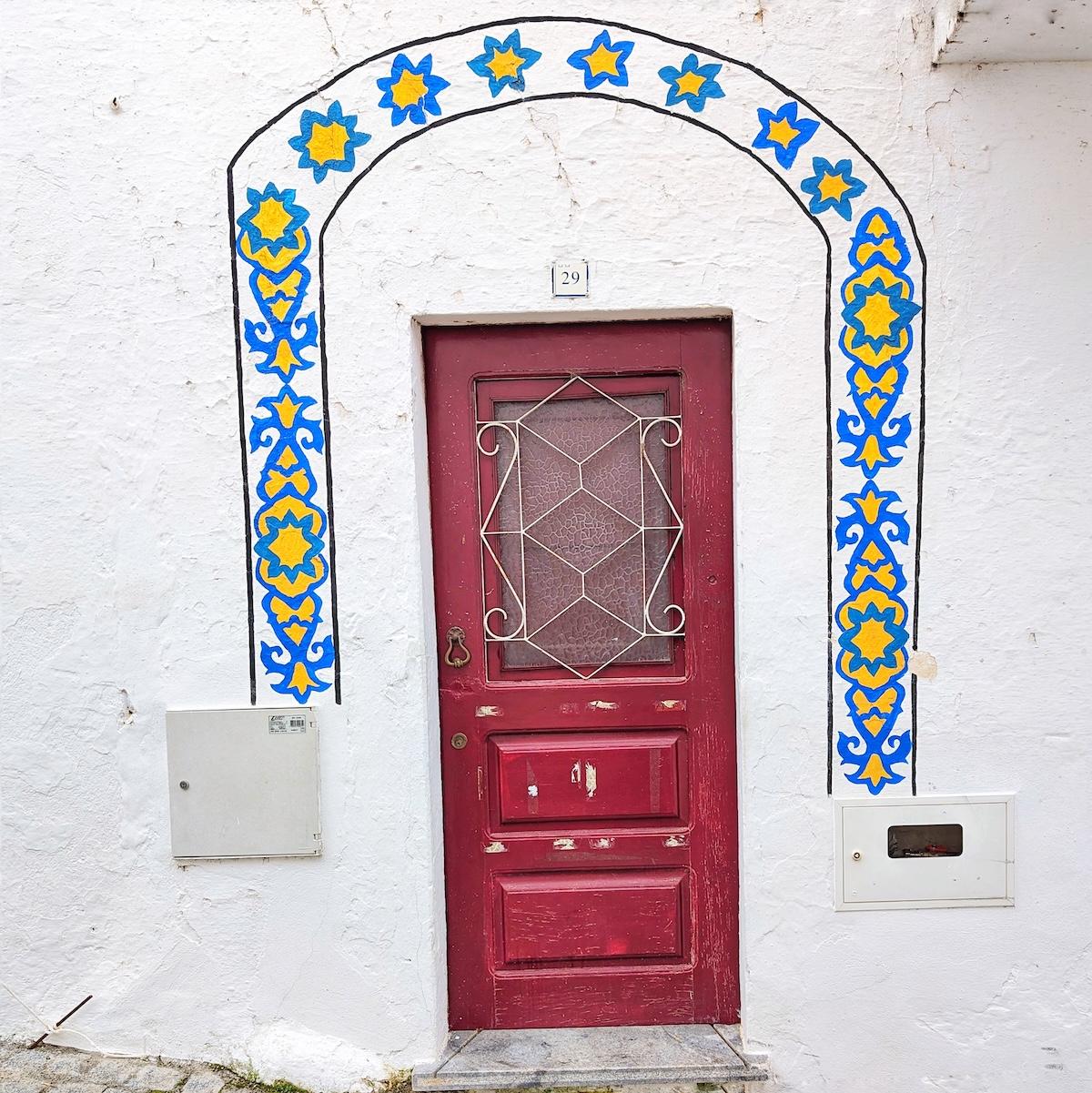 A weathered burgundy door featuring a white metal grate, framed by a hand-painted blue and yellow floral star arch on a white wall.