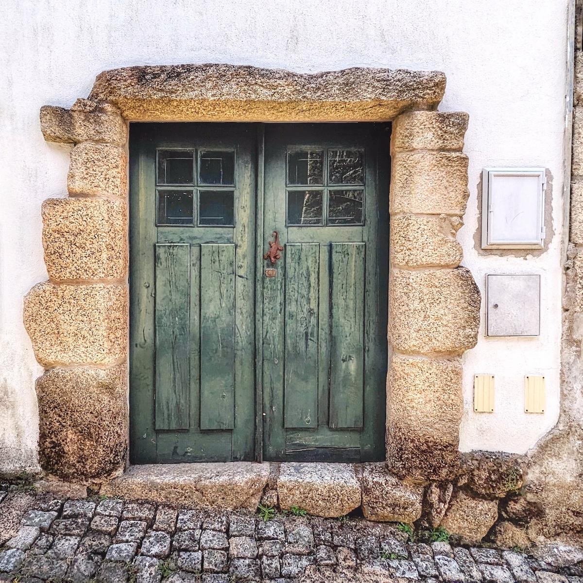 A rustic, dark green wooden double door set into a massive, uneven hand-cut stone archway on a cobblestone street.