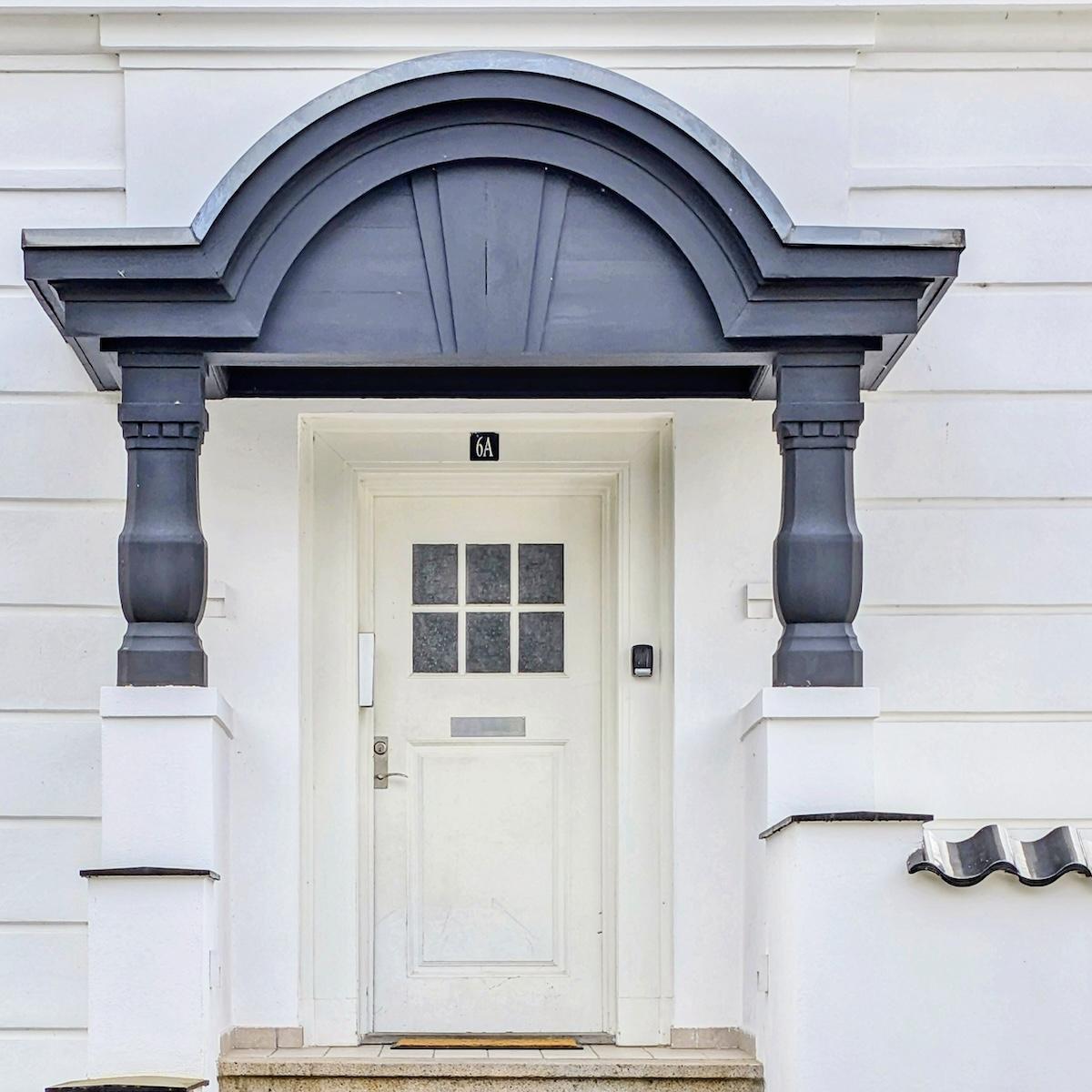 A clean white door with a six-pane window, framed by a heavy, dark charcoal-grey portico supported by two thick columns. The surrounding building is white with horizontal architectural lines.