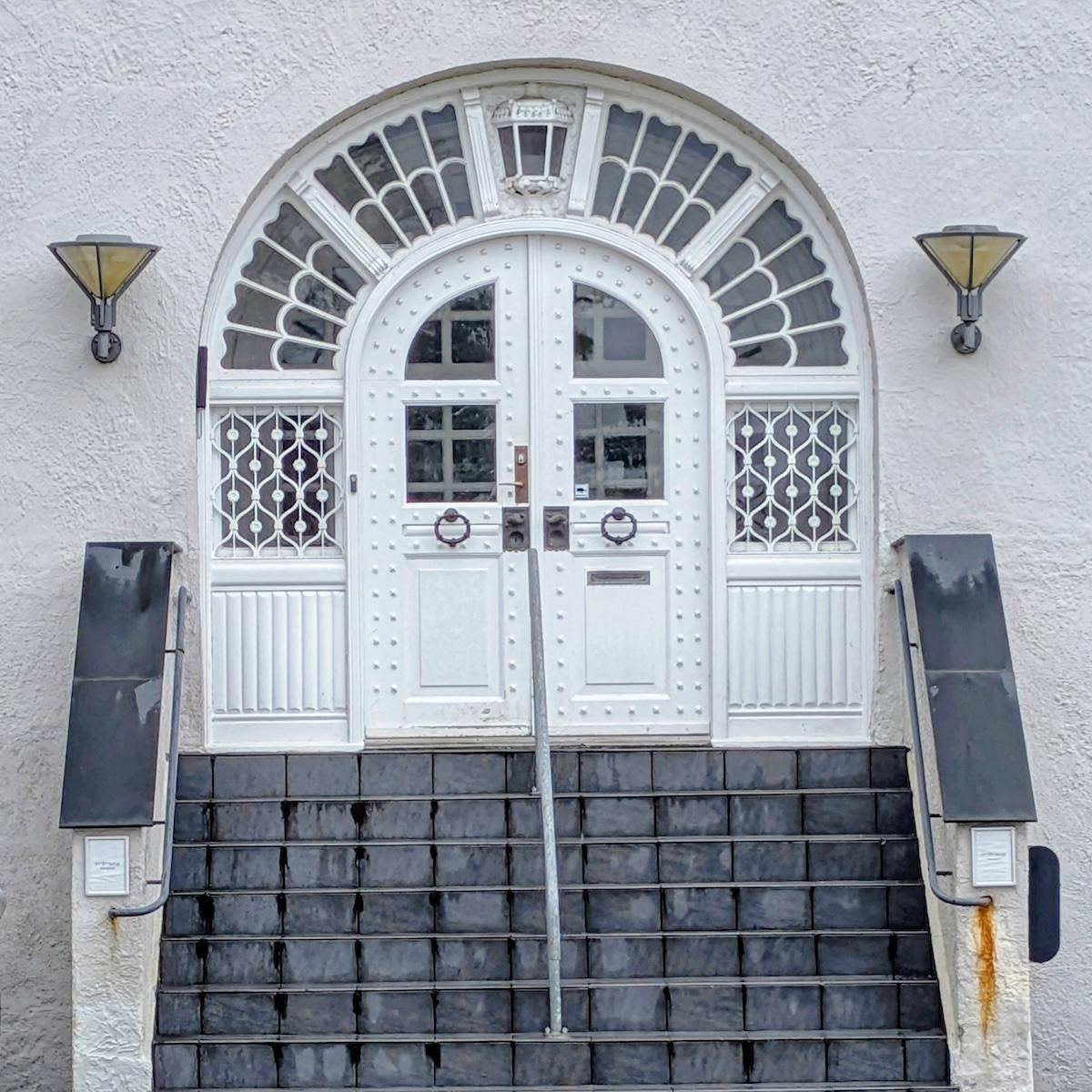 Symmetrical white double doors featuring a riveted design and dark circular knockers. The doors are set beneath an expansive arched transom window with decorative white wooden slats.