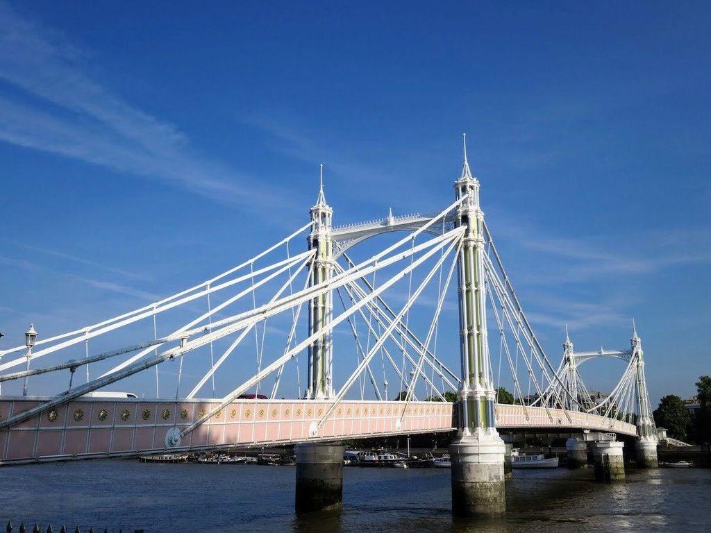 A side profile of the Albert Bridge, a bright suspension bridge painted in pastel shades of pink, blue, and white. The intricate ironwork and ornate towers stand out against a deep blue sky, with the River Thames flowing calmly beneath it.