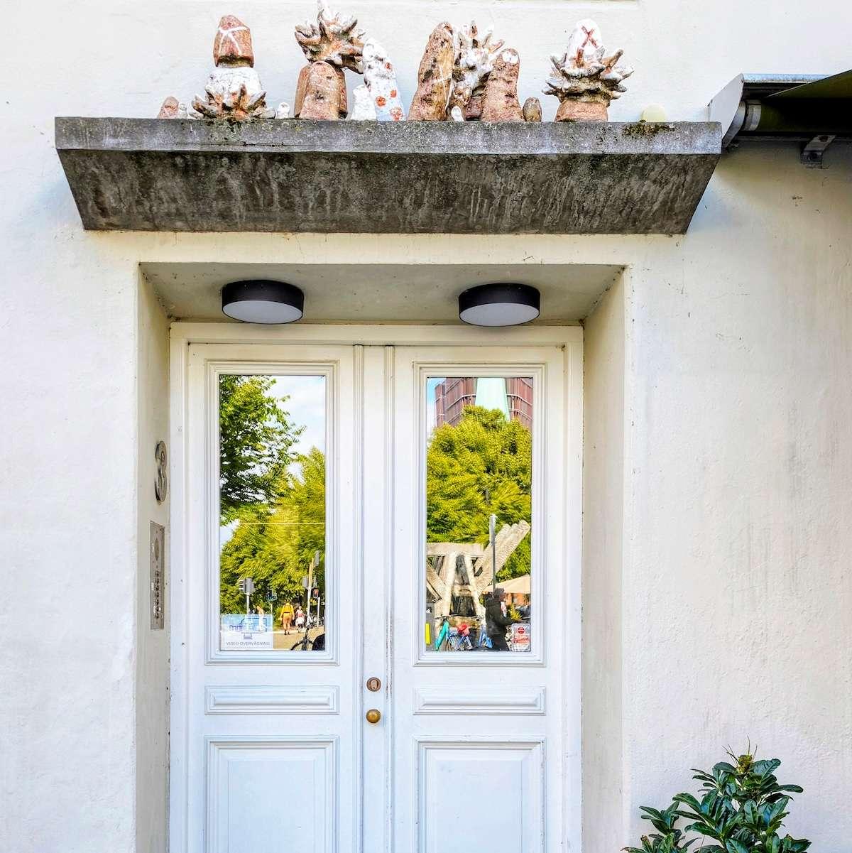 A set of white wooden double doors with large glass panes that reflect a green park. Above the concrete canopy sits a collection of unique, hand-crafted stone sculptures.
