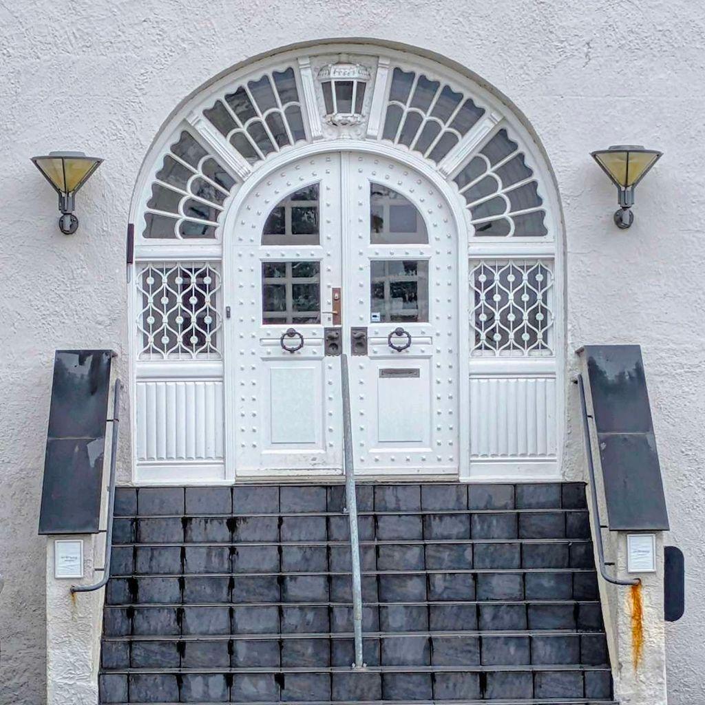 Symmetrical white double doors featuring a riveted design and dark circular knockers. The doors are set beneath an expansive arched transom window with decorative white wooden slats.