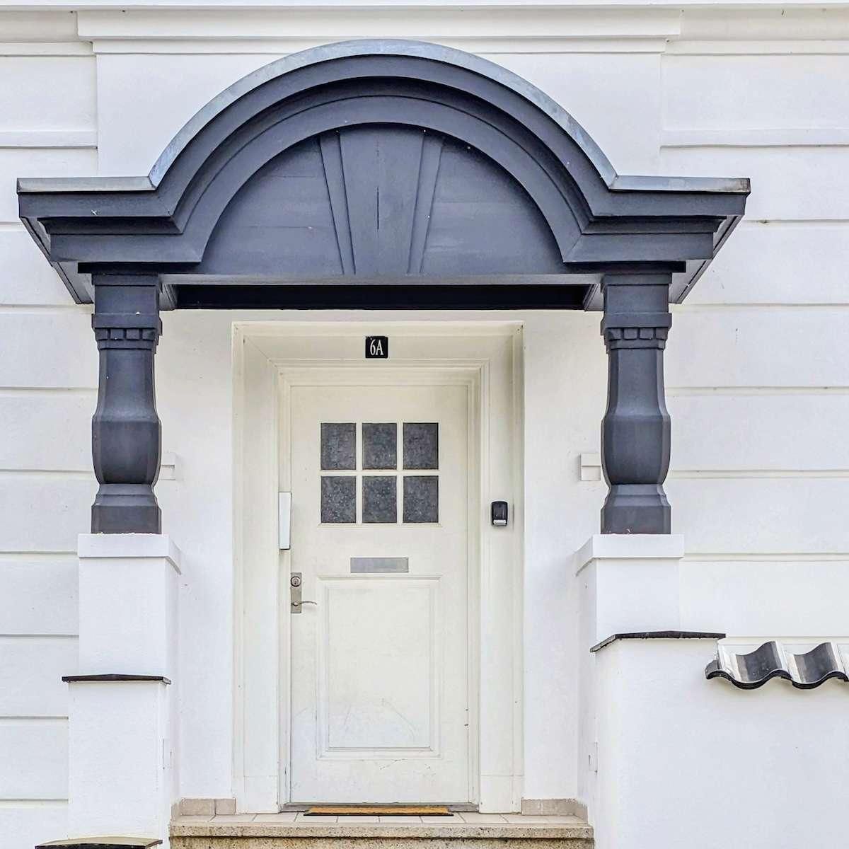 A clean white door with a six-pane window, framed by a heavy, dark charcoal-grey portico supported by two thick columns. The surrounding building is white with horizontal architectural lines.