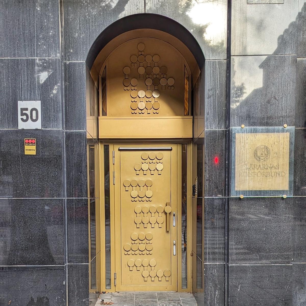 A striking gold-colored metal door and transom decorated with a series of raised circular geometric patterns. The door is recessed into a sleek, dark grey polished stone facade.