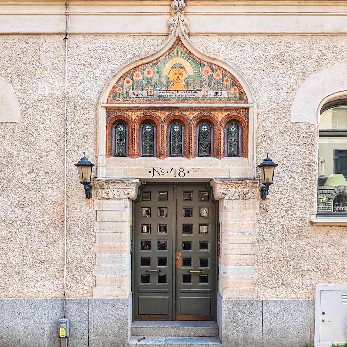 A dark olive-green door with small square window panes, positioned beneath an elaborate arched mural. The mural features a central figure with a halo, floral motifs, and the date 1899.