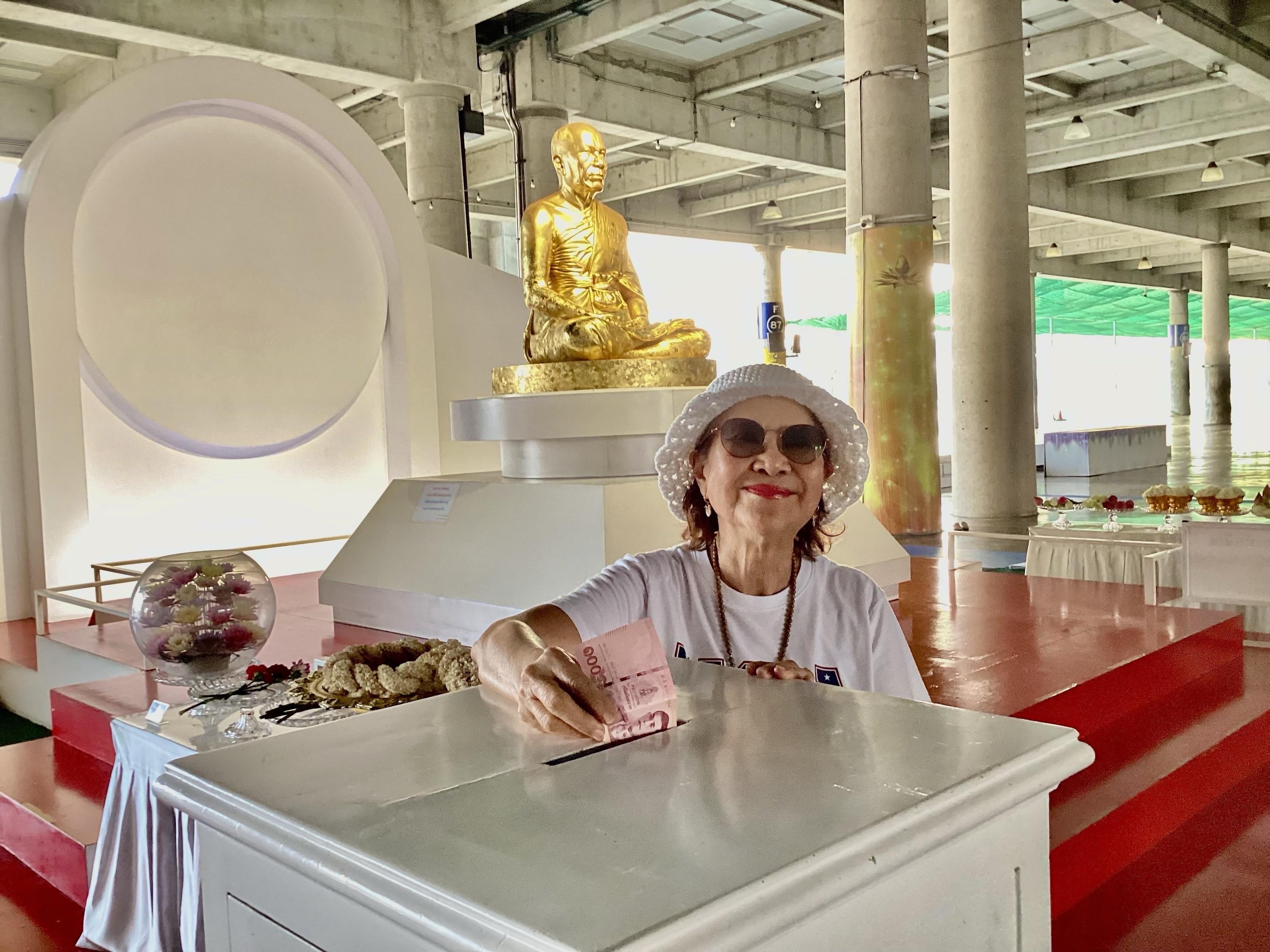 A woman wearing a white hat and sunglasses is placing money into a donation box in a temple setting. In the background, there is a golden seated Buddha statue and various decorative offerings. The space is brightly lit with a modern architectural design.