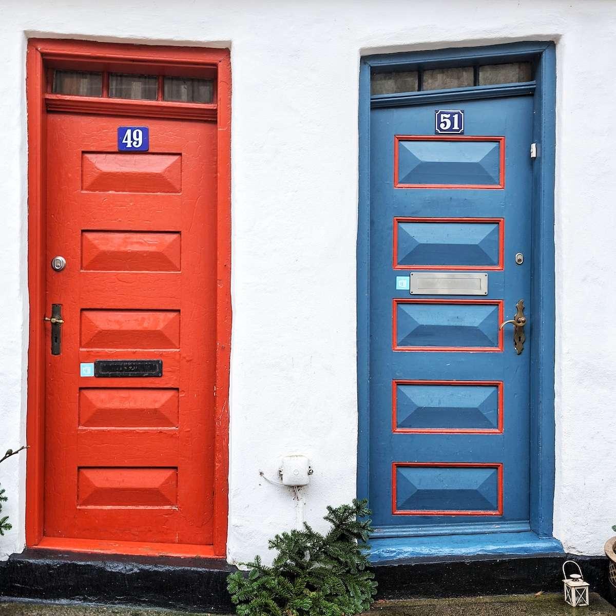 Two narrow, vertical-paneled wooden doors—one bright red and one blue with red accents—standing side-by-side in a white stucco wall. The doors are slightly slanted, reflecting the character of an older building.