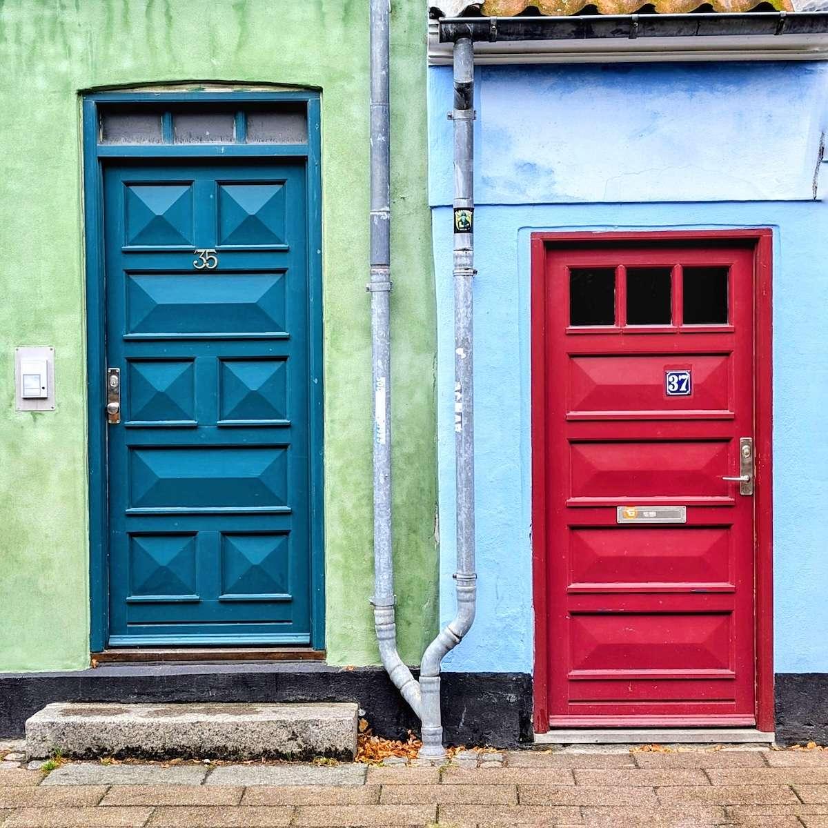 A teal blue door with raised geometric panels sits next to a bright red door on a building with lime green and sky blue stucco walls. A grey metal downspout runs vertically between the two colorful entrances.