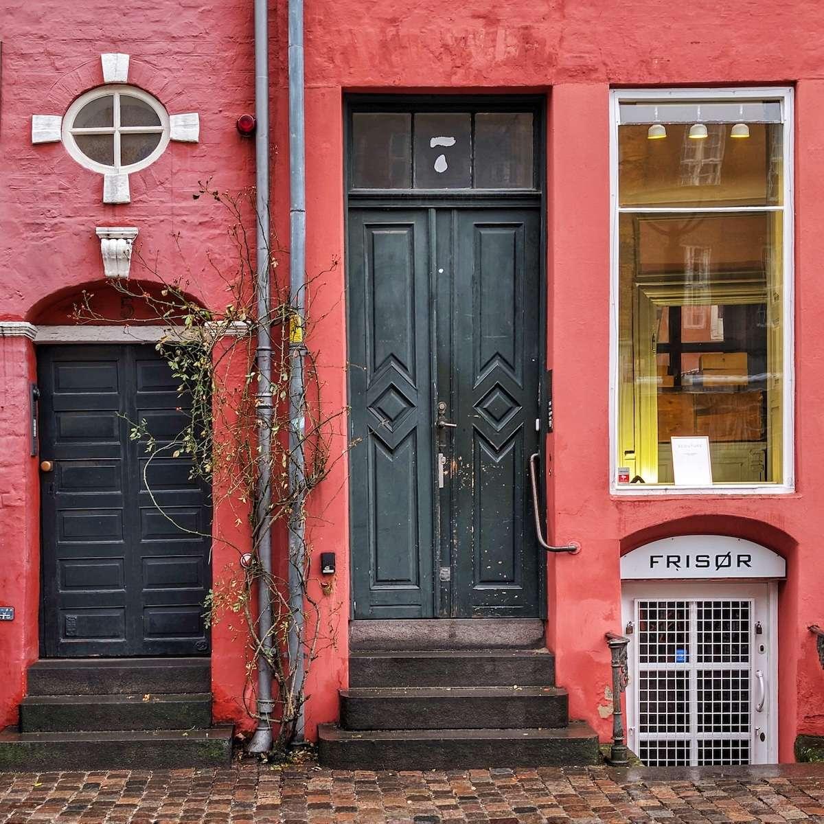 Two dark green or black wooden doors of different heights set into a textured, reddish-pink wall. The taller central door features a diamond-patterned panel, while a small white door for a "Frisør" (barber) is nestled into the basement level.