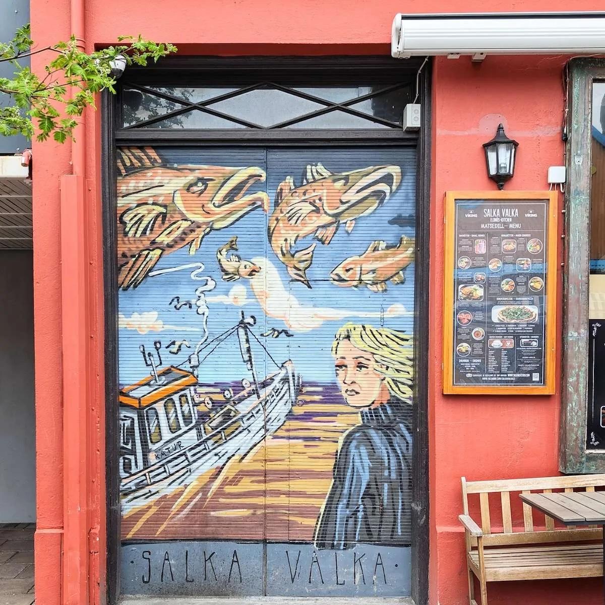 A metal roll-down security door painted with a detailed mural of large fish, a fishing boat, and a person looking out to sea. The door is part of the "Salka Valka" restaurant in Iceland.