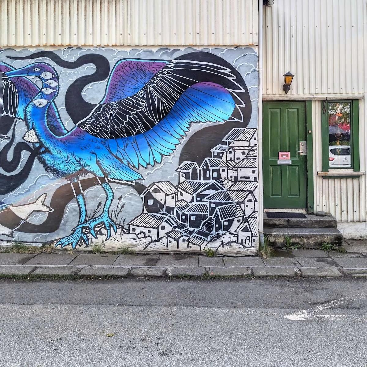 A simple green wooden door set into a white corrugated metal building. The adjacent wall features a massive, colorful mural of a blue heron standing over a stylized village of black-and-white houses.