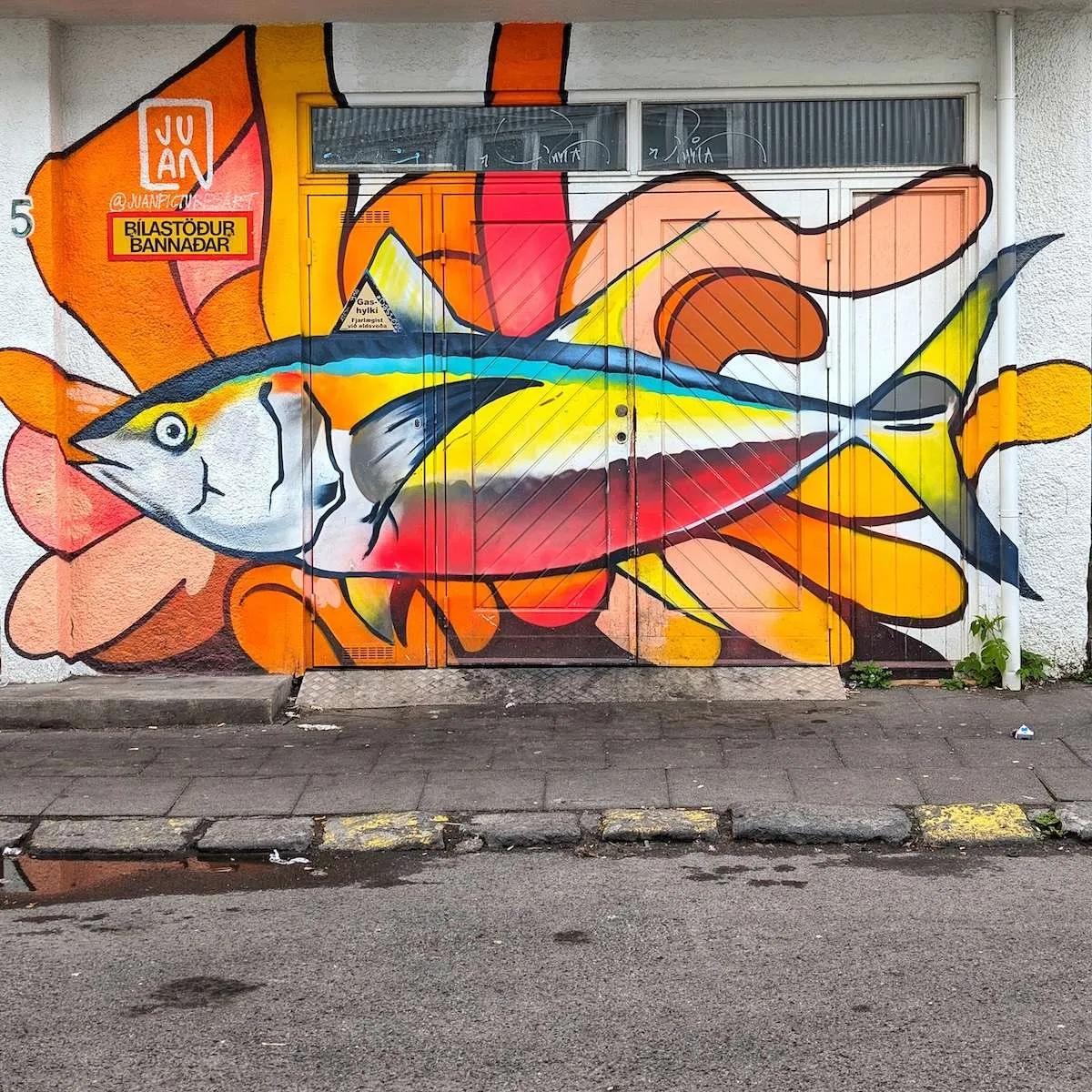 A wide garage-style entrance covered in a bold, colorful mural of a giant tuna fish against a bright orange and yellow background. The art spans across multiple door panels on a white textured building.