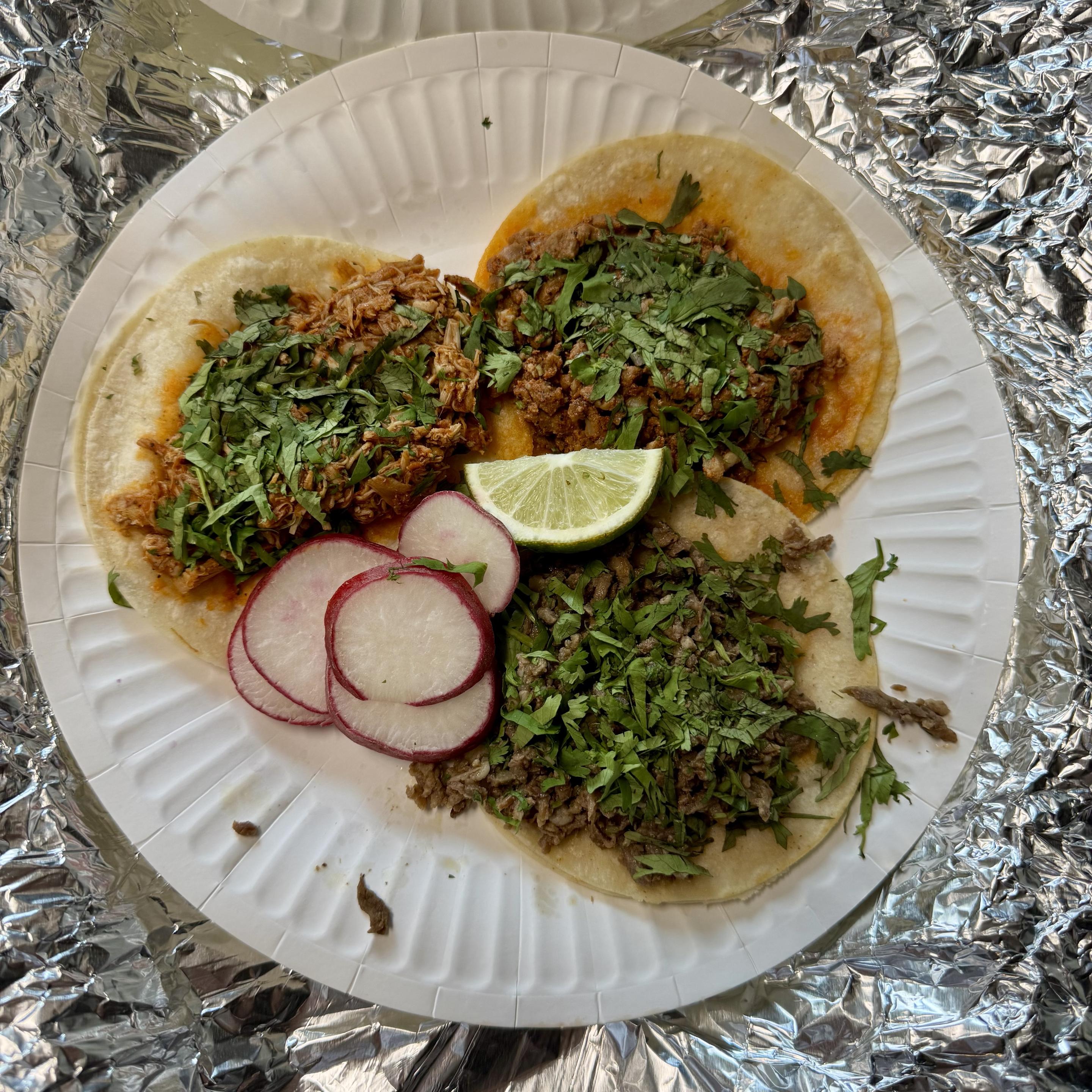 A plate with three tacos topped with chopped cilantro. One taco contains shredded meat, another features ground meat, and a third has a mix of both. Slices of radish and a lime wedge are included. The plate is set on foil.