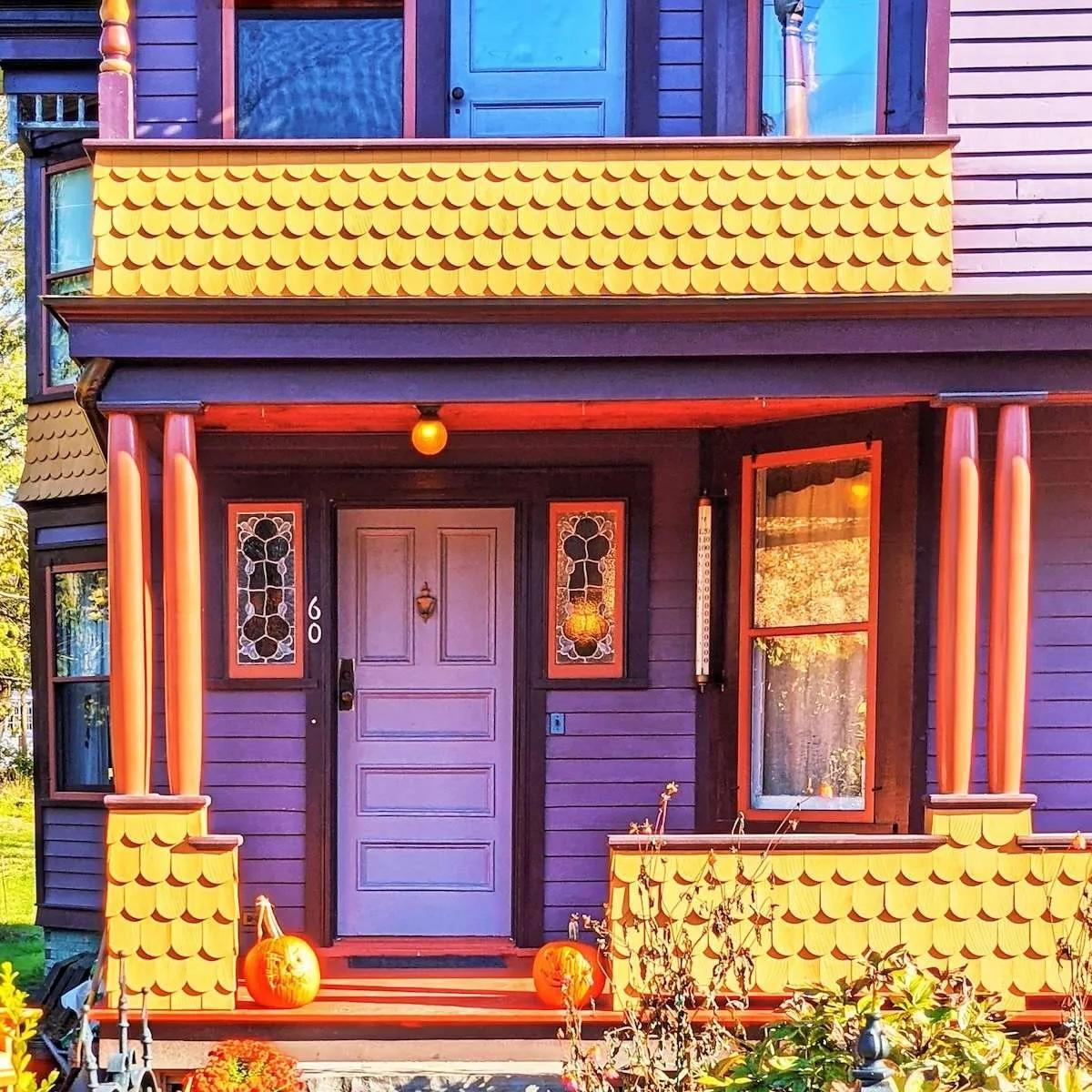 A light lavender door set against a dark purple wooden house with bright orange trim. The porch features yellow fish-scale shingles and two pumpkins, creating a colorful, autumnal Victorian aesthetic.