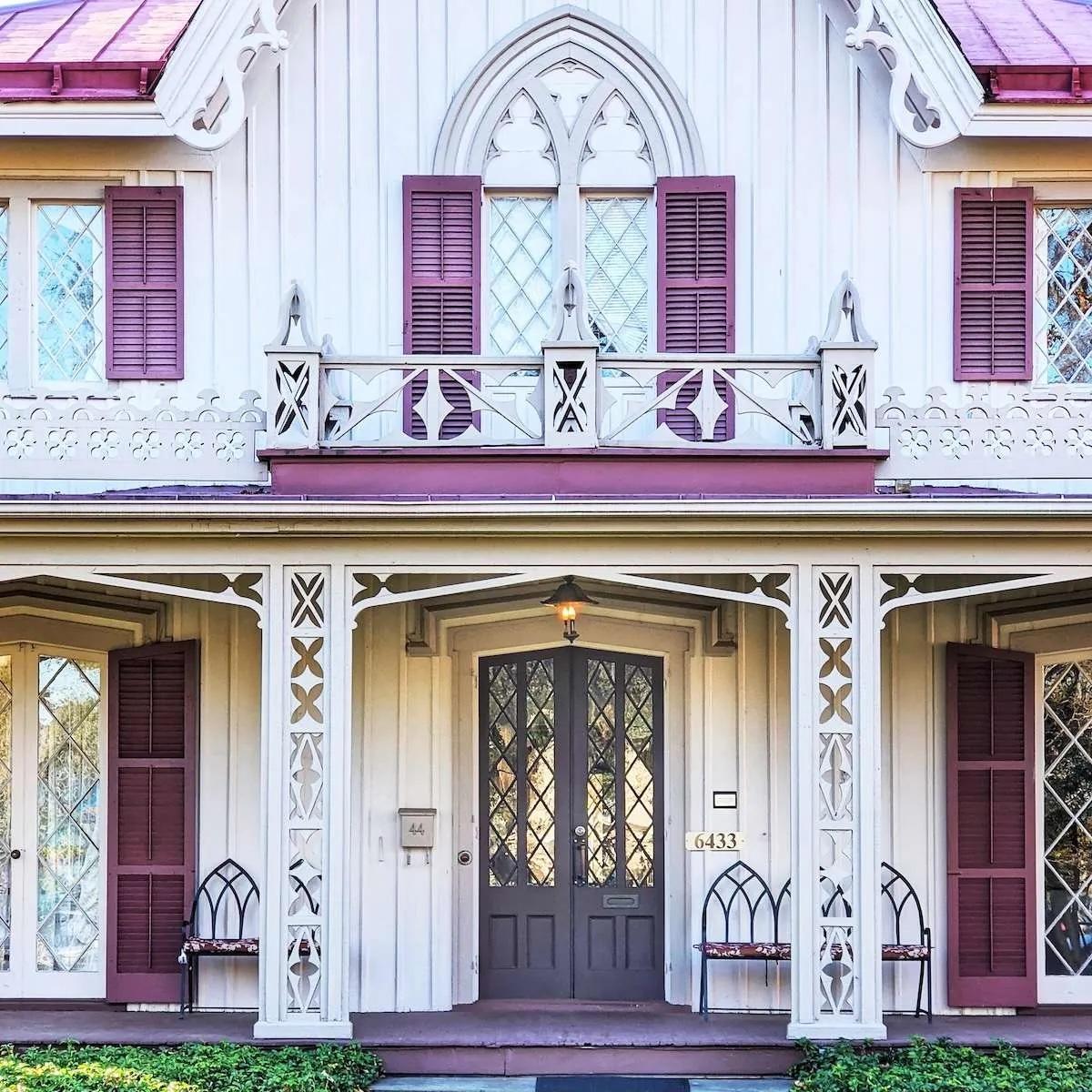 A pair of dark grey-purple doors with diamond-paned glass, situated on a white Gothic Revival house with decorative "gingerbread" trim. The entrance is flanked by purple shutters and white latticework porch supports.