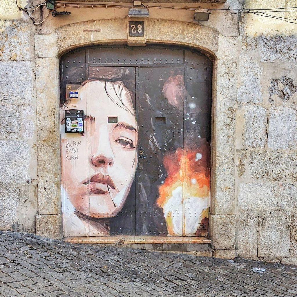 A wide metal door on a cobblestone street featuring a large-scale portrait of a woman with a flame effect, accompanied by the hand-painted text "BURN BABY BURN."