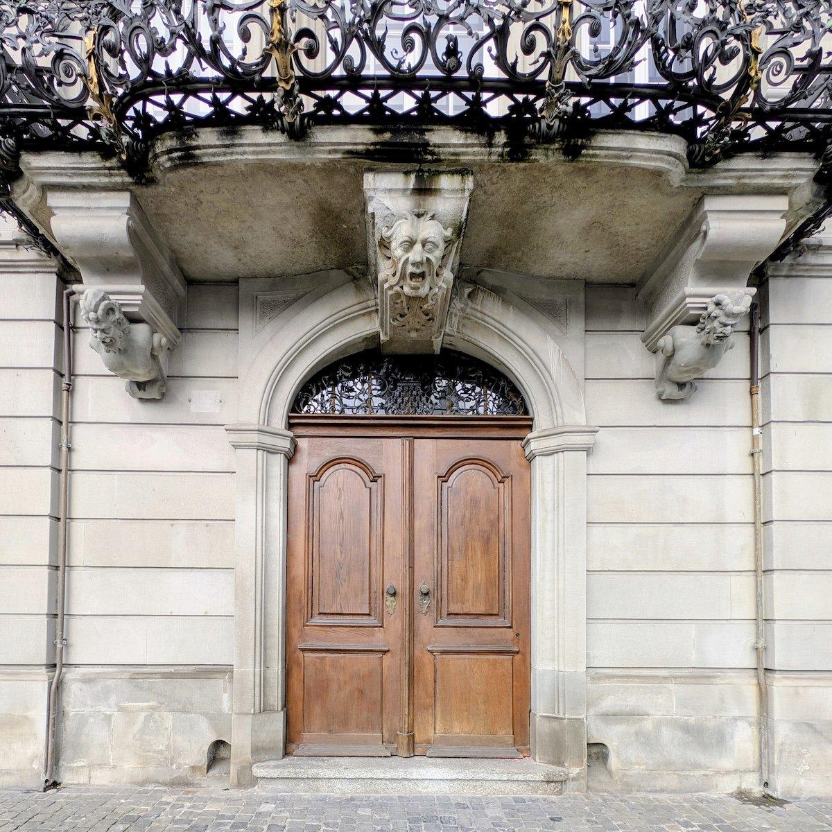 Elegant arched wooden double doors set beneath a stone balcony supported by three expressive gargoyle-like mascaron carvings, with an ornate wrought iron railing above.