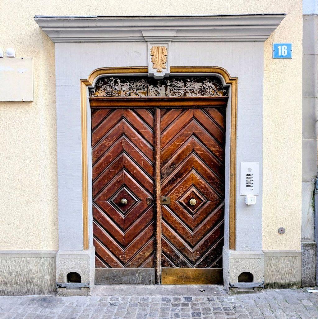 Historic wooden double doors in Zurich featuring a bold chevron and diamond parquet design, framed by a grey stone portal with gold accents and a delicate ironwork transom.