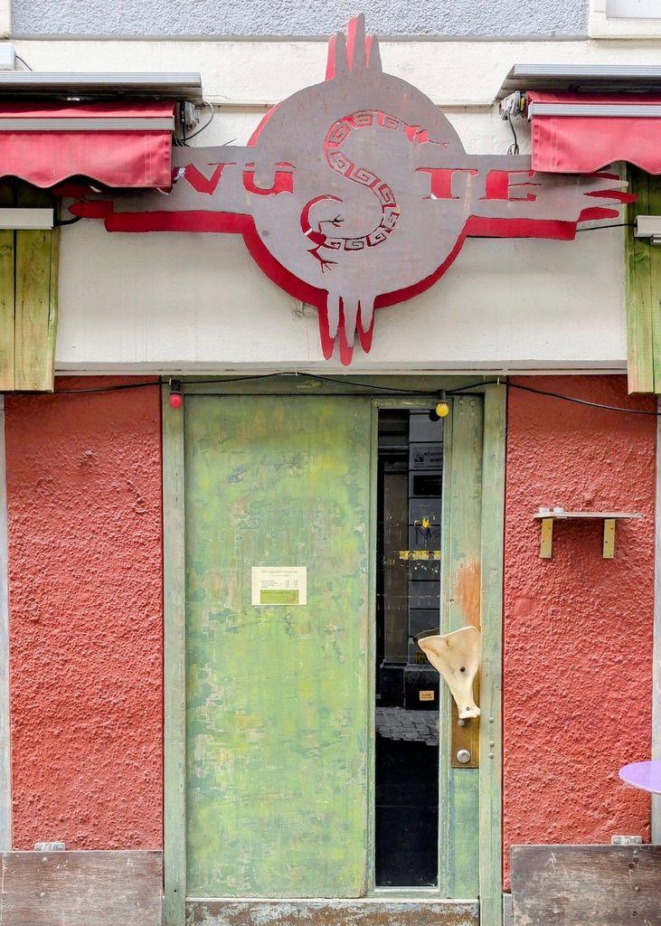 A weathered, lime-green door with a long vertical glass pane and a bacalhau-shaped handle, topped by a large industrial metal sign featuring a red snake and the word "WÜSTE."
