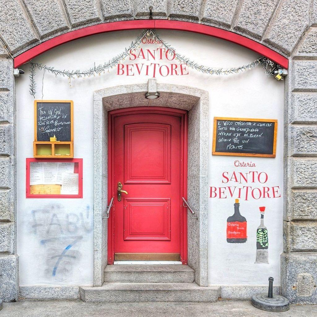 Bright red door serving as the entrance to Osteria Santo Bevitore in Zurich, surrounded by white walls with hand-painted wine bottles and chalkboard menus.