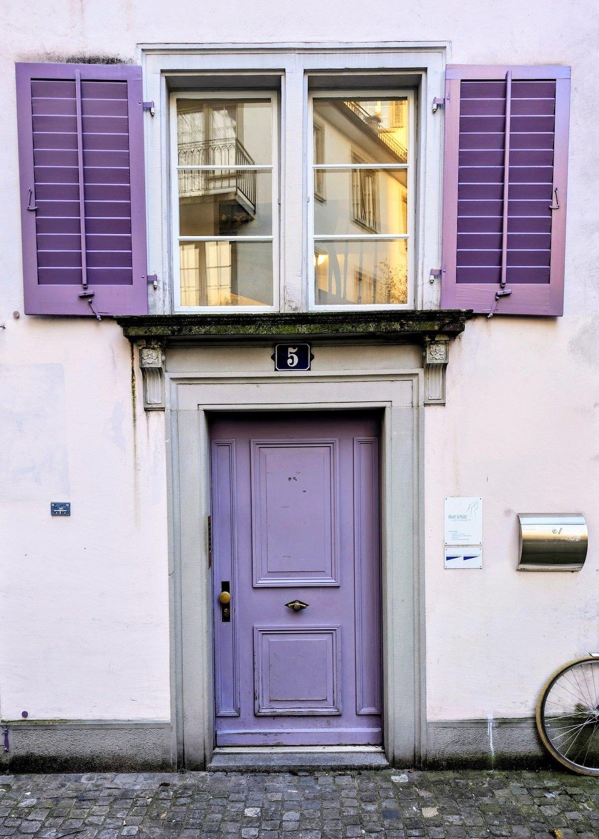 A classic lavender-purple paneled door in Zurich set within a grey stone frame, topped by matching purple shutters and a transom window reflecting neighboring buildings.
