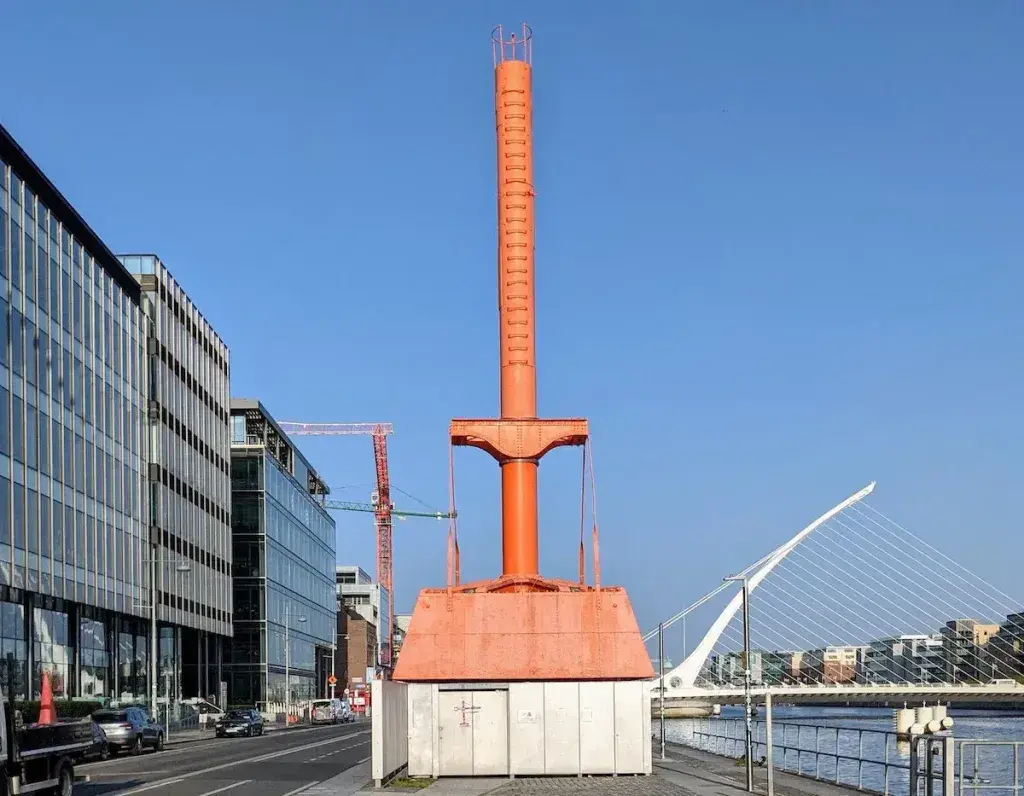 The Diving Bell: Tall orange buoy-like tower mounted on a rectangular base along a riverside promenade, framed by modern glass office buildings to the left and a white cable-stayed bridge spanning the river in the background.