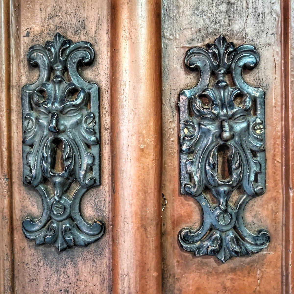 Two ornate matching metal keyhole plates shaped as stylized grotesque faces with open mouths and scrolling details, mounted on a worn wooden double door showing aged patina.