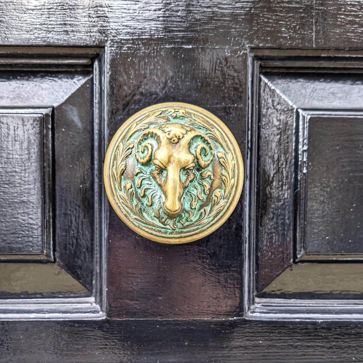 Ornate round brass door knob featuring a ram's head surrounded by leaf motifs and green verdigris patina, mounted on a glossy black paneled front door.