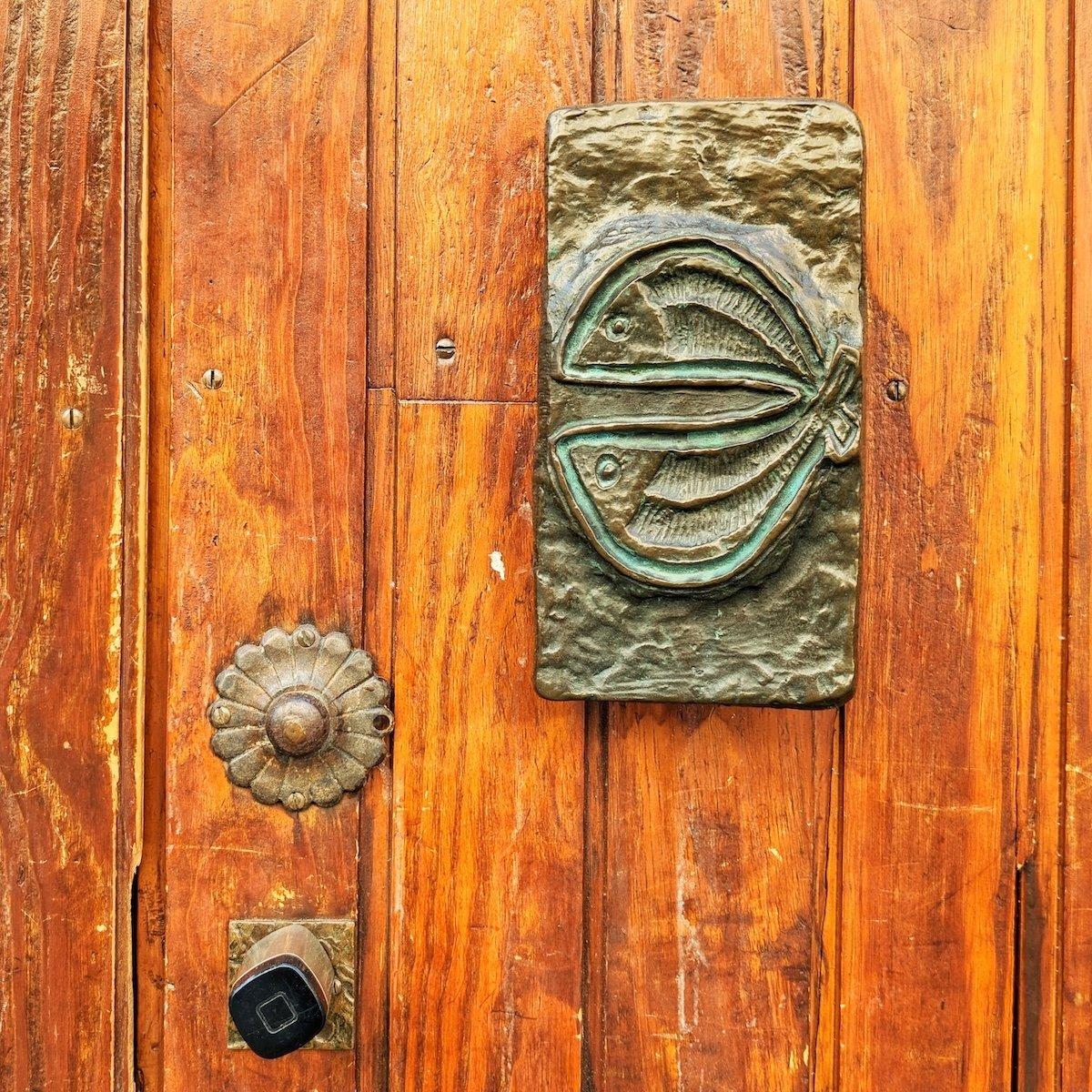 Bronze rectangular door plate embossed with a stylized pair of fish mounted on a weathered wooden door, accompanied by an antique flower-shaped knob and a modern square deadbolt.