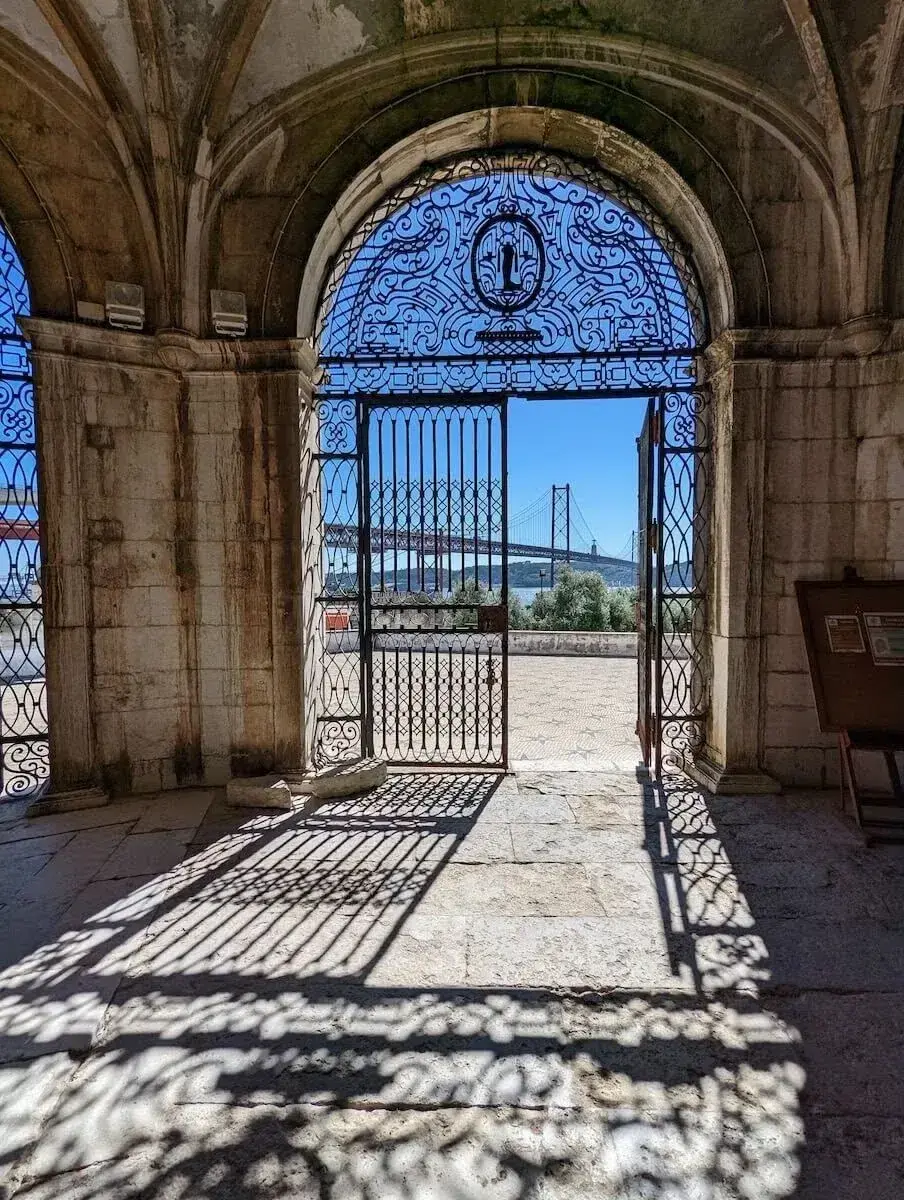 Ornate arched iron gate in a weathered stone arcade framing a distant suspension bridge over the river, sunlight casting decorative lattice shadows across the stone floor.